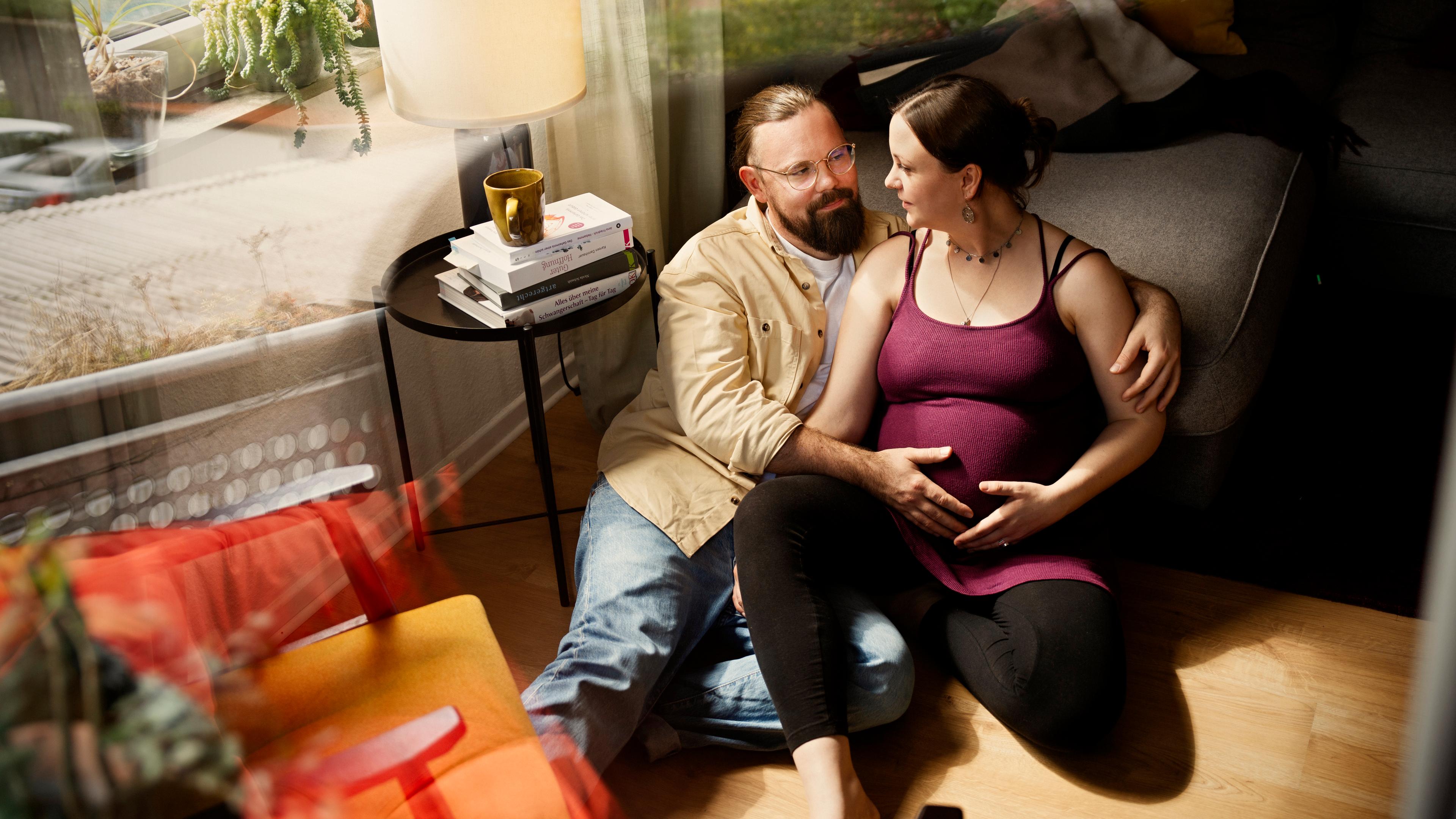 Gitta und Julian sitzen im Wohnzimmer auf dem Boden und schauen sich an. Er hat seine Hand auf ihren Bauch gelegt.