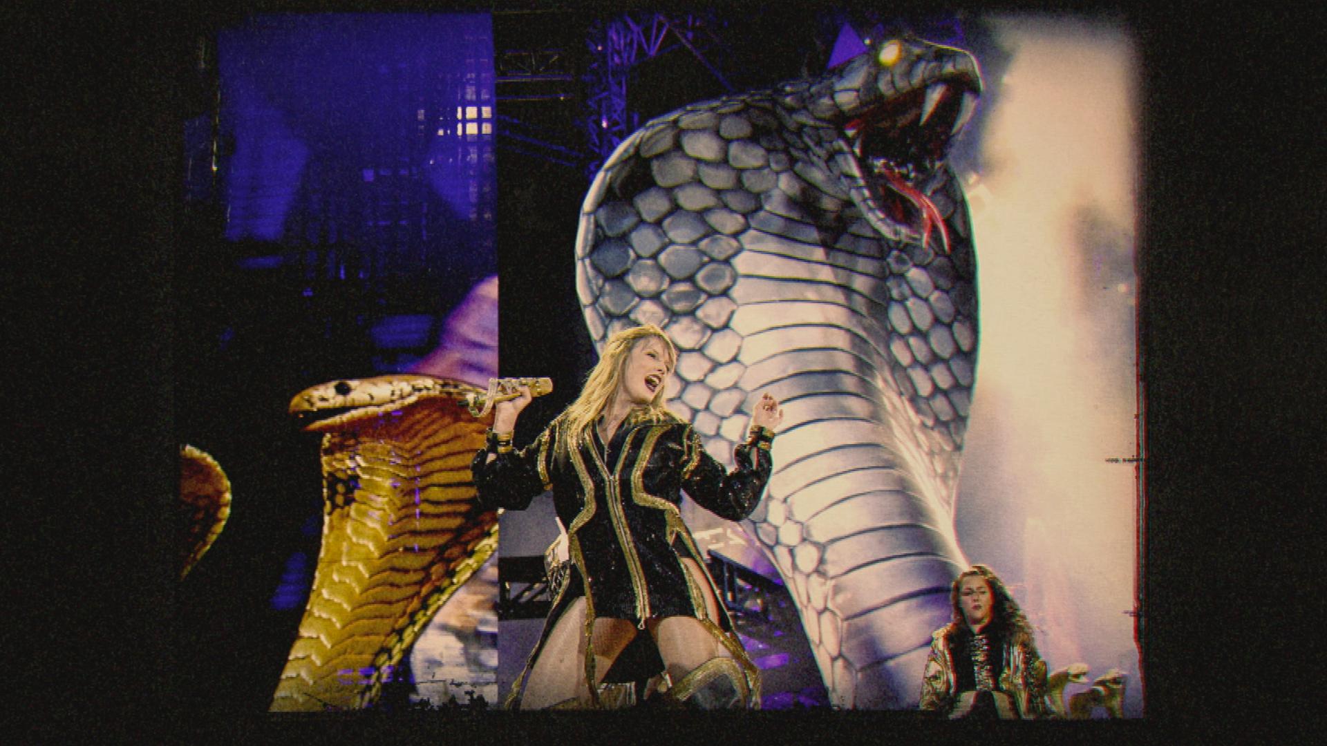 Taylor Swift steht in einem schwarzen, goldverzierten Bühnenkostüm vor zwei großen, aufgerichteten Schlangenfiguren auf einer Bühne.