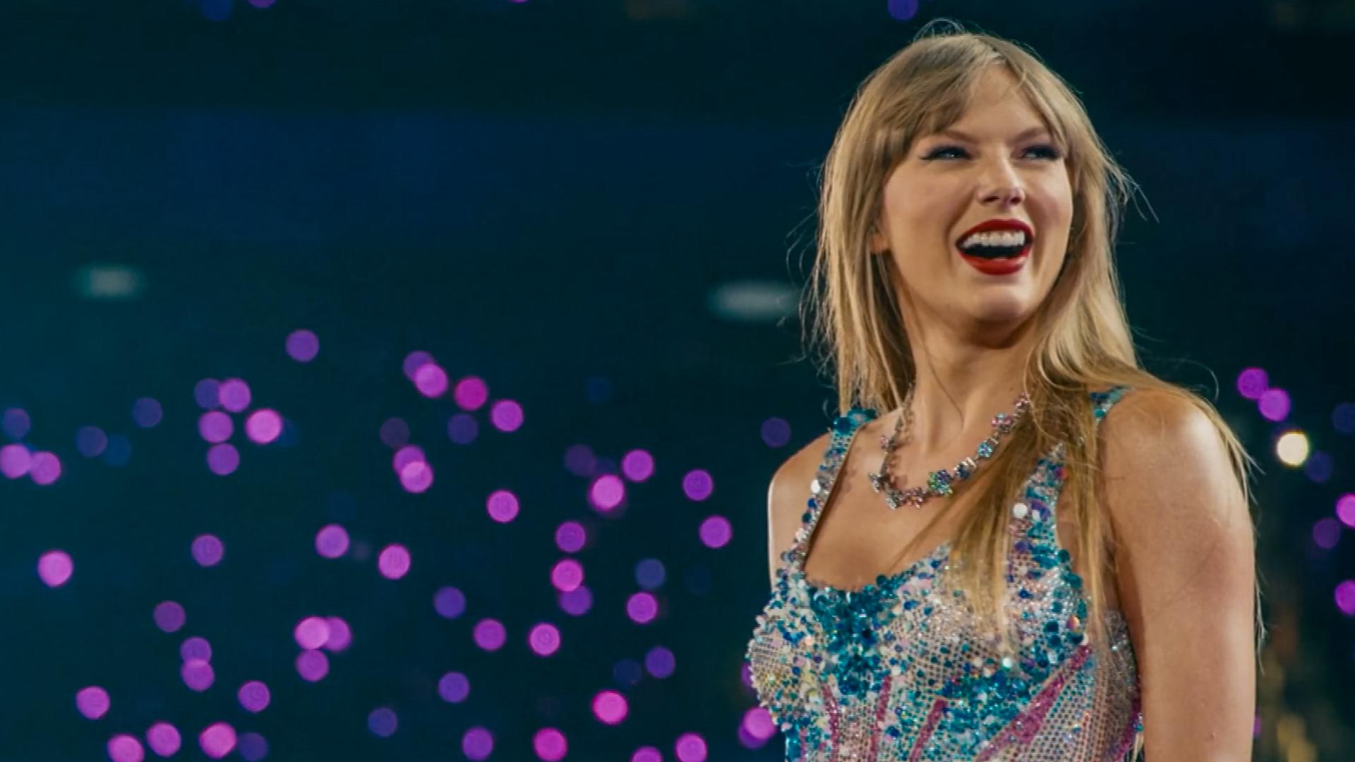 Taylor Swift steht auf der Bühne und trägt ein glitzerndes, buntes Kleid und eine auffällige Halskette vor unscharfem Hintergrund mit lila Lichtpunkten.