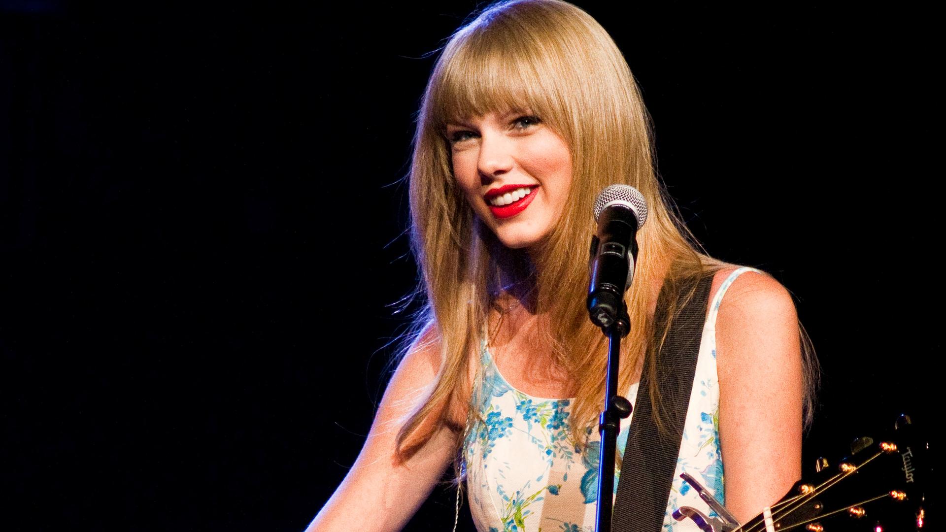 Taylor Swift steht auf der Bühne und trägt ein glitzerndes, buntes Kleid und eine auffällige Halskette vor unscharfem Hintergrund mit lila Lichtpunkten.