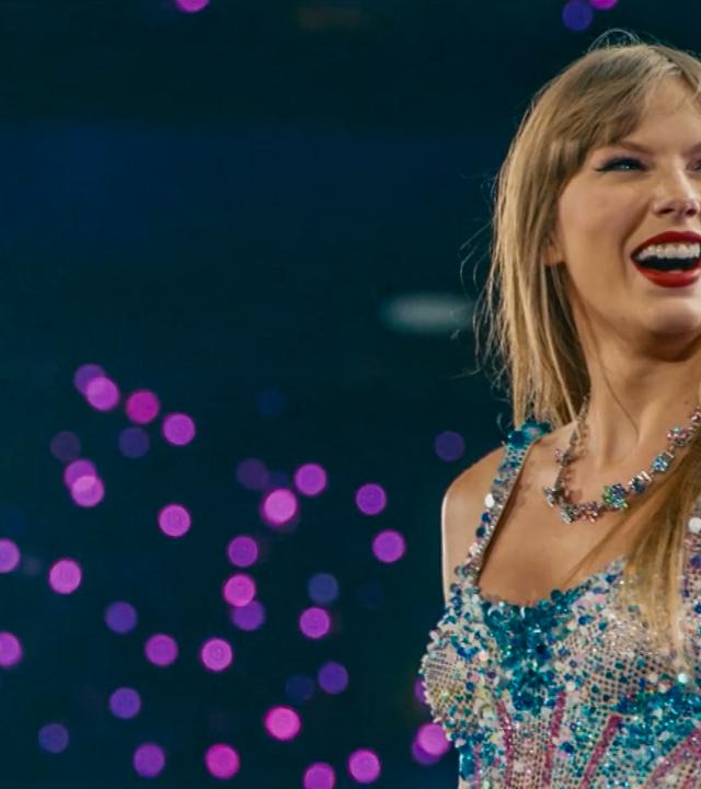 Taylor Swift steht auf der Bühne und trägt ein glitzerndes, buntes Kleid und eine auffällige Halskette vor unscharfem Hintergrund mit lila Lichtpunkten.