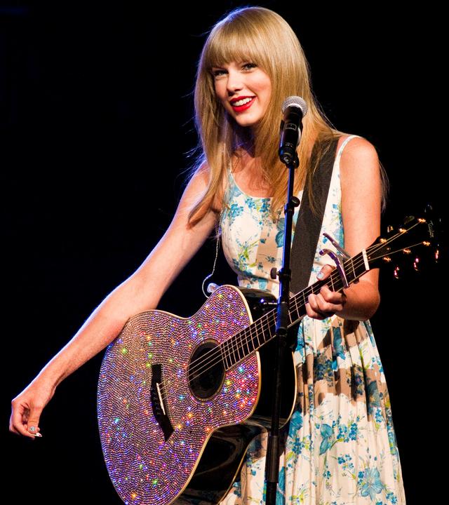 Taylor Swift steht auf der Bühne und trägt ein glitzerndes, buntes Kleid und eine auffällige Halskette vor unscharfem Hintergrund mit lila Lichtpunkten.