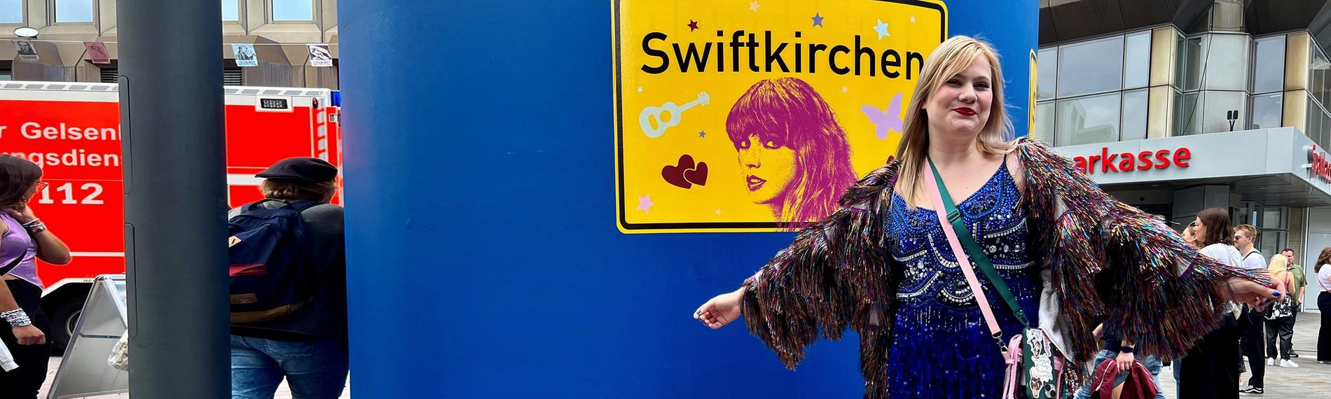 Kim steht in einer Fußgängerzone vor einer blauen Litfaßsäule, darauf ist ein großes gelbes Schild mit der Aufschrift "Swiftkirchen". Sie trägt ein kurzes blaues Kleid und blaue Stiefel.