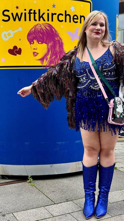 Kim steht in einer Fußgängerzone vor einer blauen Litfaßsäule, darauf ist ein großes gelbes Schild mit der Aufschrift "Swiftkirchen". Sie trägt ein kurzes blaues Kleid und blaue Stiefel.