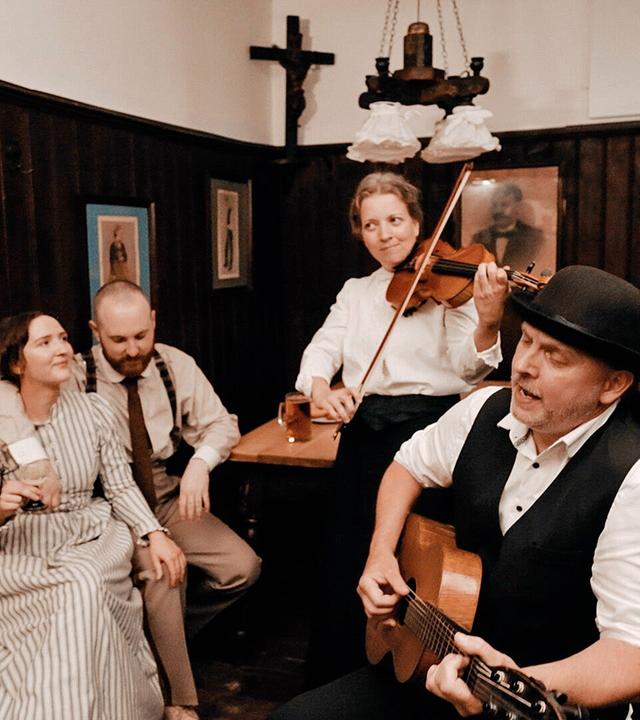 Musikanten mit Geige, Gitarre und Ziehharmonika vor  Wirthaus-Publikum