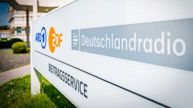 Beitragsservice von ARD, ZDF und Deutschlandradio: Weißes Außenschild mit den Logos von ARD (blau), ZDF (orange) und Deutschlandradio sowie der Aufschrift „Beitragsservice“. Das Schild steht vor einem modernen Gebäude, im Vordergrund ist eine Grünfläche mit Gras sichtbar.