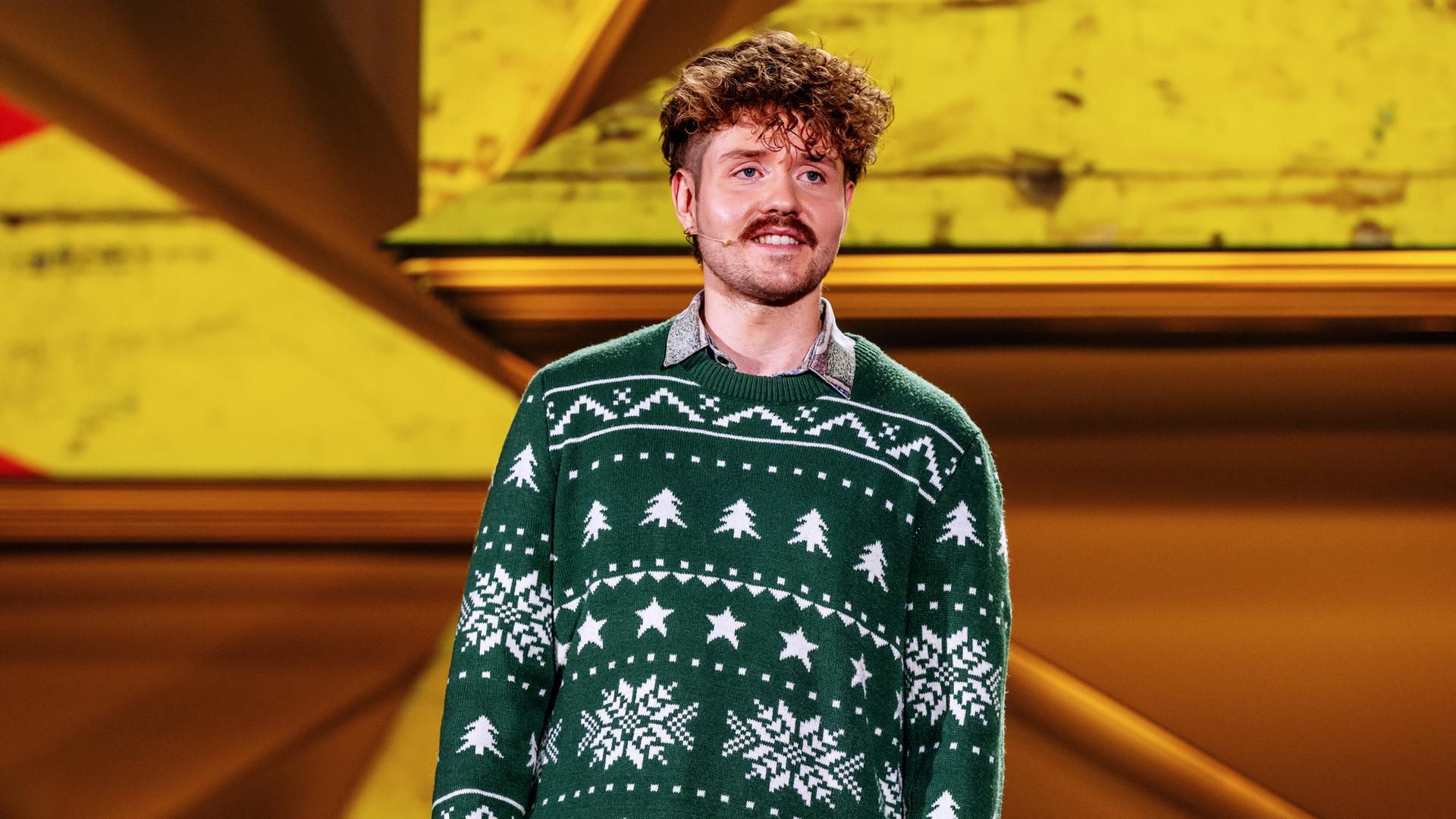 Benedikt Mitmannsgruber steht in der Mitte des Bildes. Der Hintergrund ist gelb beleuchtet und er trägt einen grünen Pullover.  