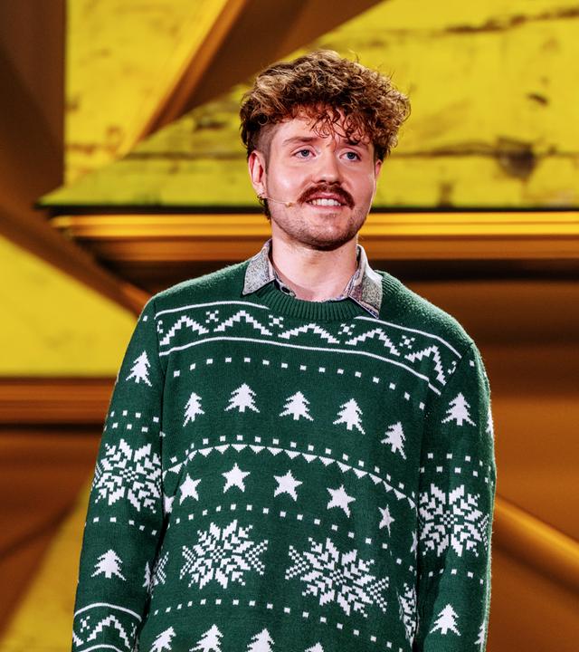 Benedikt Mitmannsgruber steht in der Mitte des Bildes. Der Hintergrund ist gelb beleuchtet und er trägt einen grünen Pullover.  