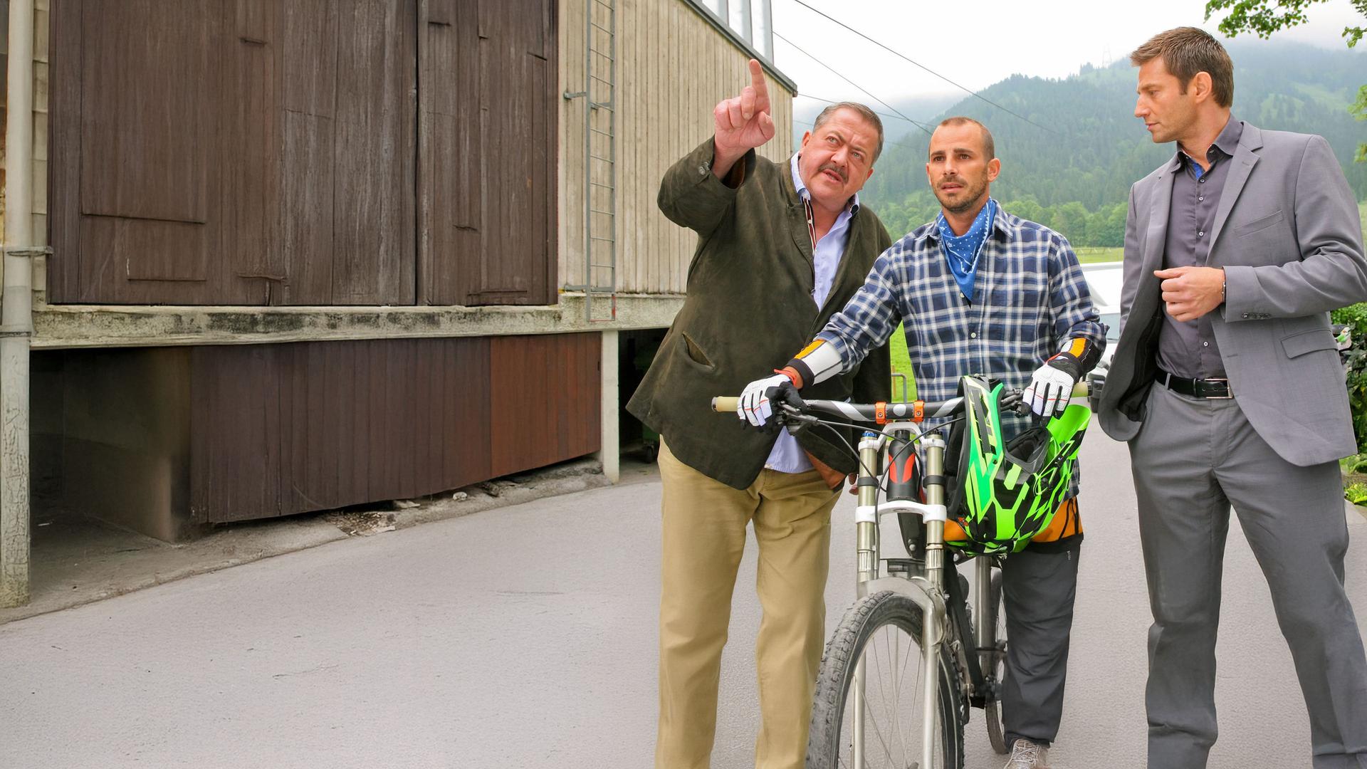 "Die Rosenheim-Cops - Bergab in den Tod": Korbinian Hofer (Joseph Hannesschläger) und Sven Hansen (Igor Jeftic) stehen an einer Talstation und befragen den Mountainbiker Peter Niemann (Maximilian Allgeier), der mit seinem Mountainbike unterwegs ist. Im Hintergrund sind bewaldete Berge zu sehen.
