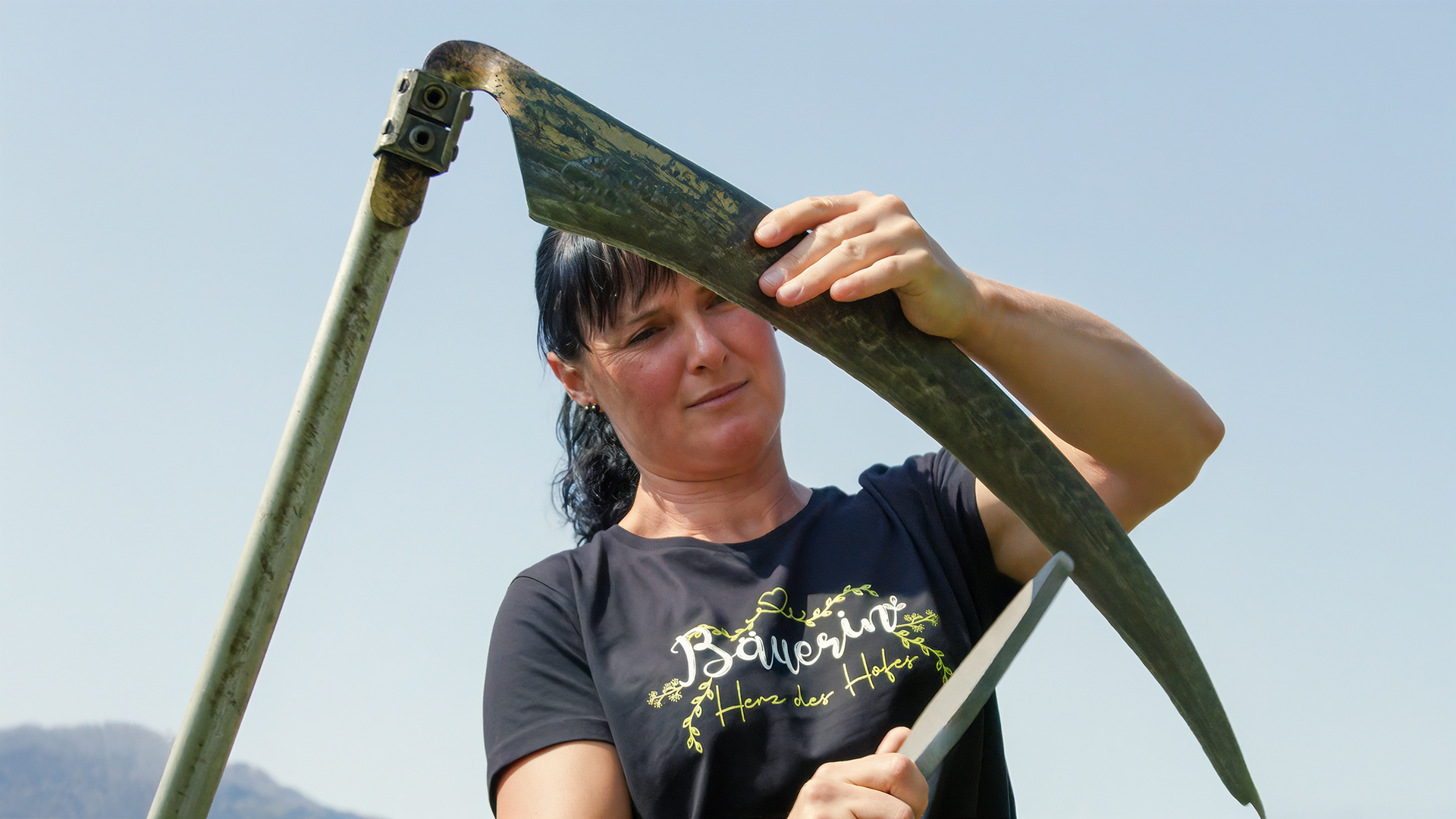 Eine Frau in einem T-Shirt mit der Aufschrift „Bäuerin“ schärft eine Sense.