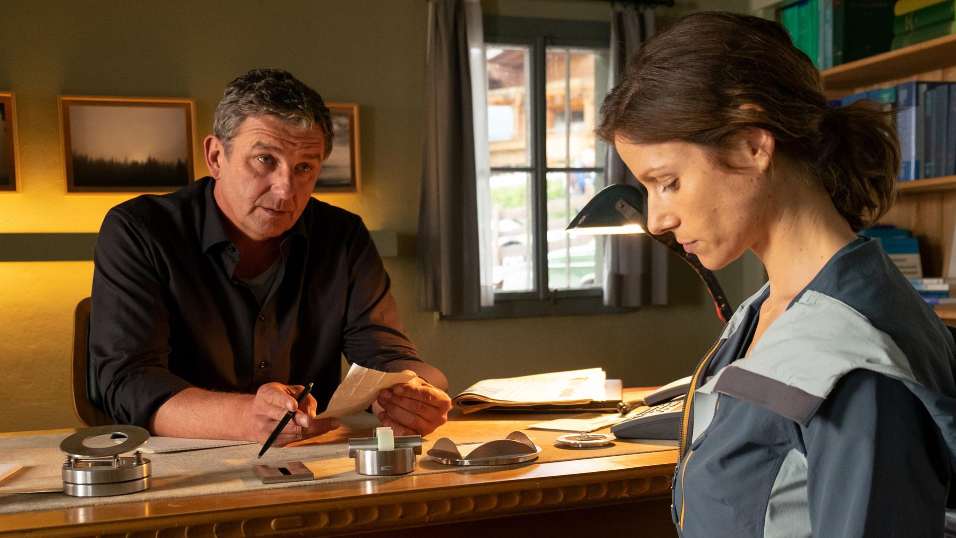 Dr. Martin Gruber (Hans Sigl) sitzt an seinem Schreibtisch in der Arztpraxis und redet auf Nima Surländer (Katrin Heß) ein, die mit gesenktem Kopf vor ihm sitzt.