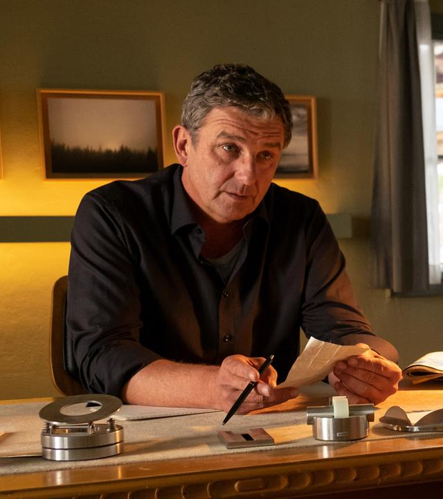 Dr. Martin Gruber (Hans Sigl) sitzt an seinem Schreibtisch in der Arztpraxis und redet auf Nima Surländer (Katrin Heß) ein, die mit gesenktem Kopf vor ihm sitzt.