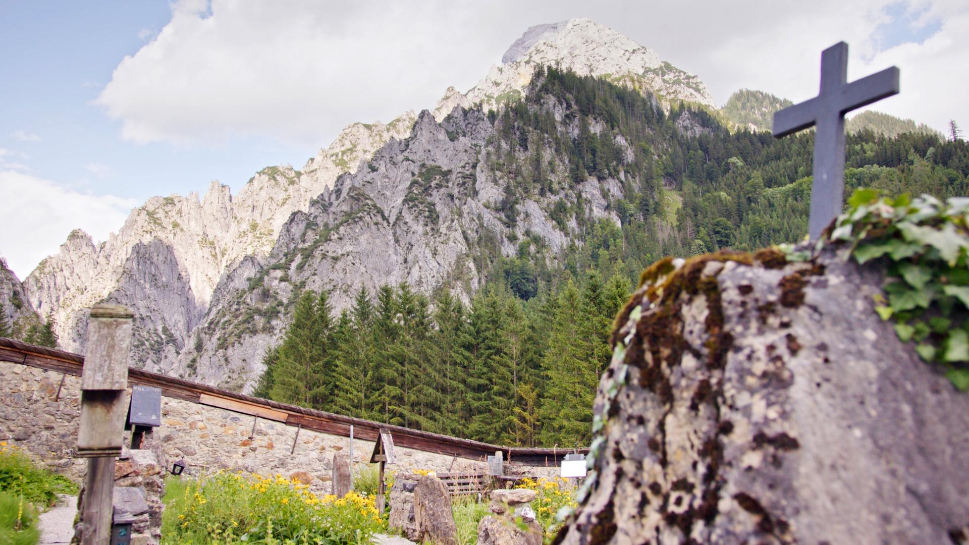 Das Bild zeigt den Bergsteigerfriedhof in Johnsbach, einem kleinen Ort im Gesäuse. Im Vordergrund ist ein Stein mit einem schwarzen Kreuz sichtbar, das auf einem moosbedeckten Felsen steht. Die Szenerie wird von der majestätischen Berglandschaft umgeben, die aus steilen, grauen Felsformationen und grünen Wäldern besteht. Im Hintergrund erheben sich hohe Berge, deren Spitzen teilweise von Wolken umhüllt sind. Der Himmel hat eine helle, bewölkte Erscheinung, und es wachsen gelbe Blumen im Vordergrund, was der Szenerie eine lebendige Note verleiht. Der Ort strahlt eine ruhige und friedliche Atmosphäre aus, die typisch für Bergregionen ist.