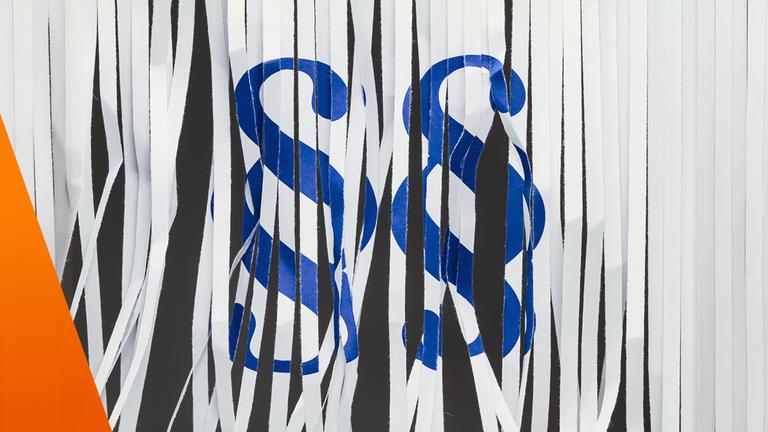 Zu sehen ist ein abstraktes Motiv aus schmalen, herabhängenden Papierstreifen, die wie geschreddert wirken. Dahinter erscheinen blaue Paragrafensymbole, die nur bruchstückhaft erkennbar sind. Der visuelle Eindruck von Zerstückelung und Überlagerung verweist auf komplexe Regelwerke, während das Thema Bürokratieabbau die Erwartung von Firmen nach Vereinfachung betont.