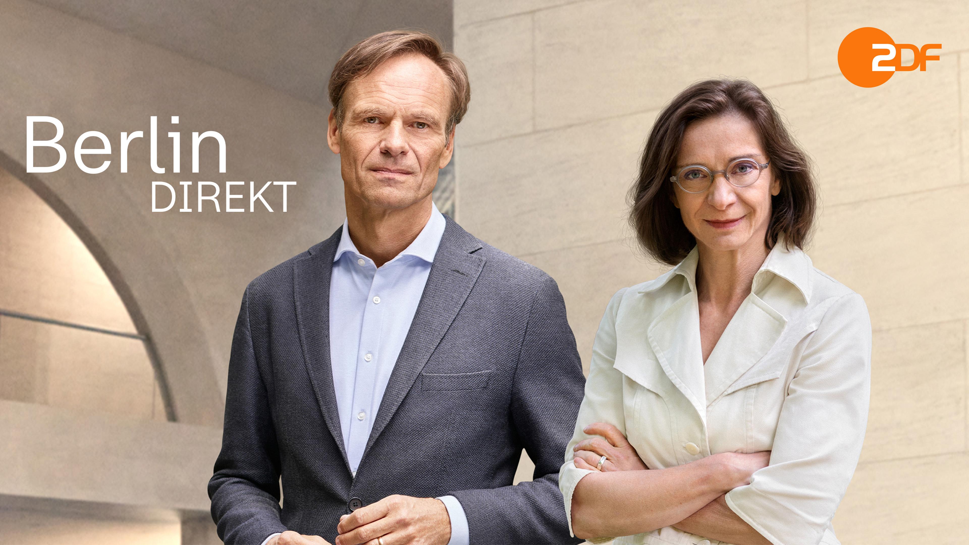 Wulf Schmiese und Diana Zimmermann stehen vor einem Regierugsgebäude in Berlin mit einer großen, runden Öffnung in der Außenmauer und blicken in die Kamera.