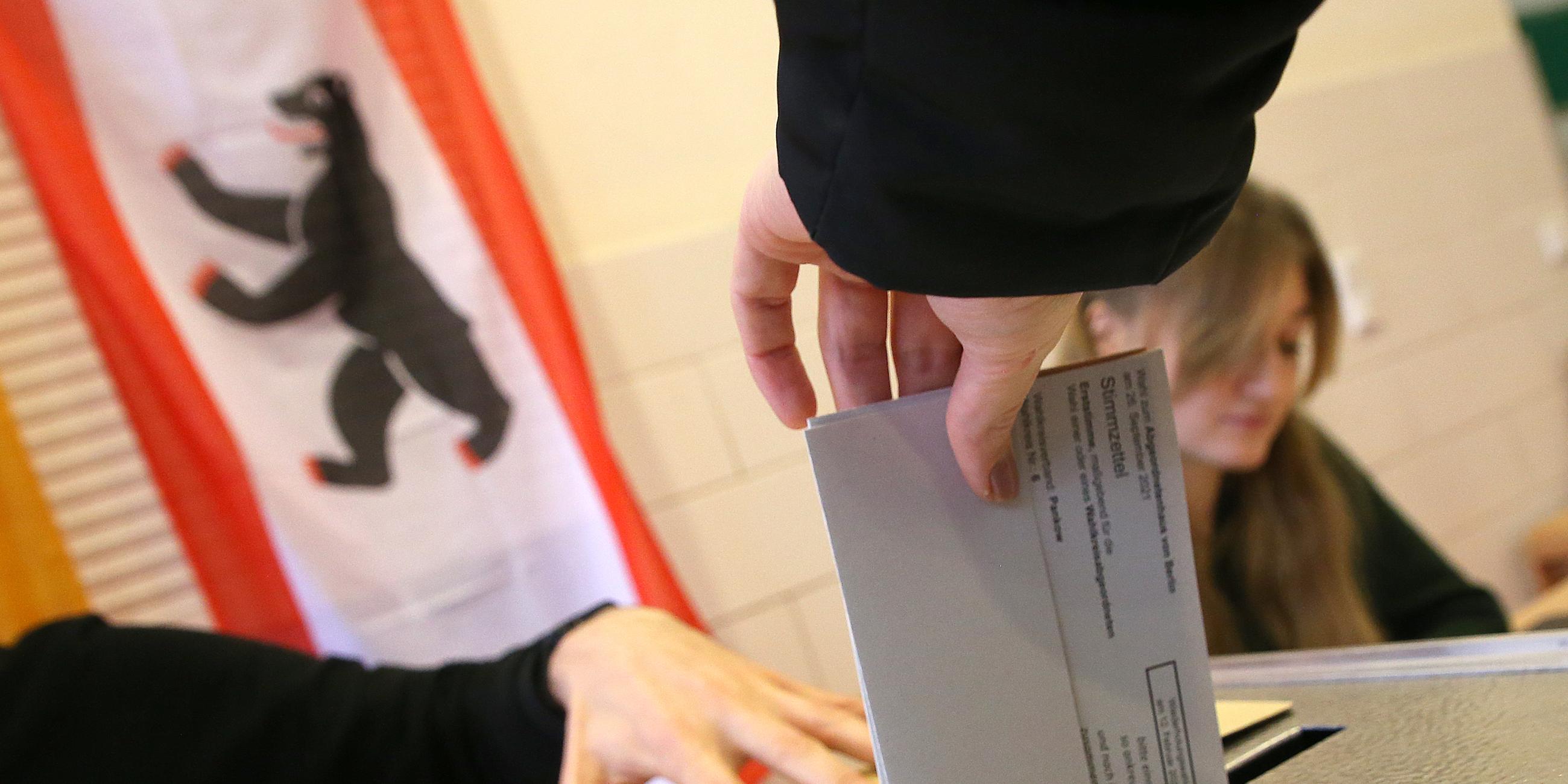 Eine Person wirft ihren Wahlzettel in die Wahlurne. Im Hintergrund sitzen zwei Wahlhelfer an einem Tisch.