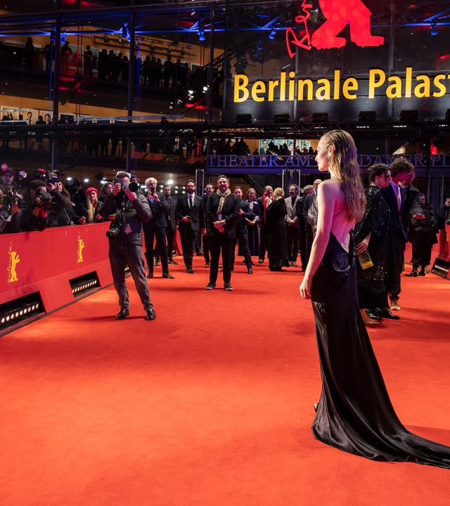 Eine Frau, die ein Abendkleid trägt, steht auf einem roten Teppich. Um sie herum befinden sich Fotografen und Publikum.