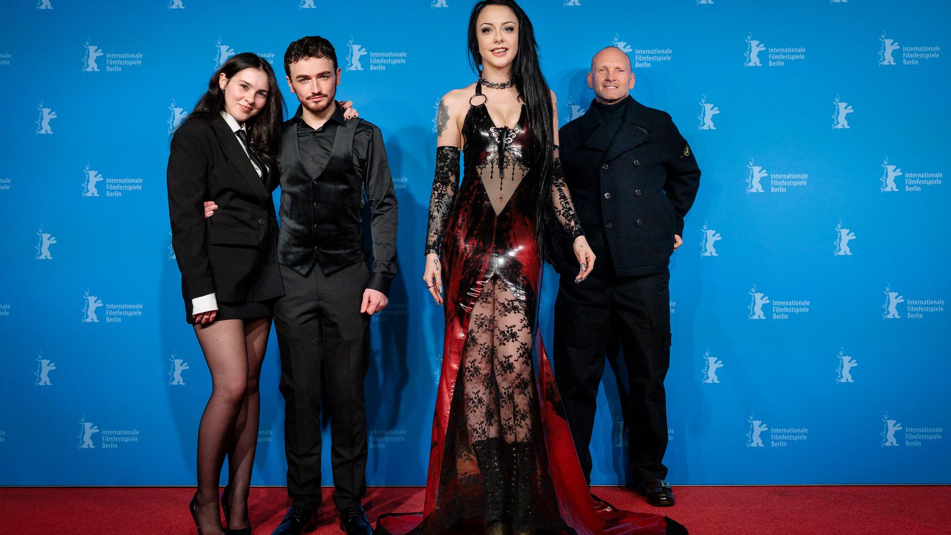 Vor dem blauen Hintergrund der Berlinale-Pressekonferenzen und auf einem roten Teppich stehen zwei Männer in schwarzer Kleidung und zwei Frauen. Eine junge Frau im Vordergrund hat lange schwarze Haare und trägt ein freizügiges Kostüm aus rotem Lack und schwarzer Spitze. Die zweite Frau trägt ein schwarzes Kostüm mit Minirock und hat den Arm um einen der Männer gelegt.