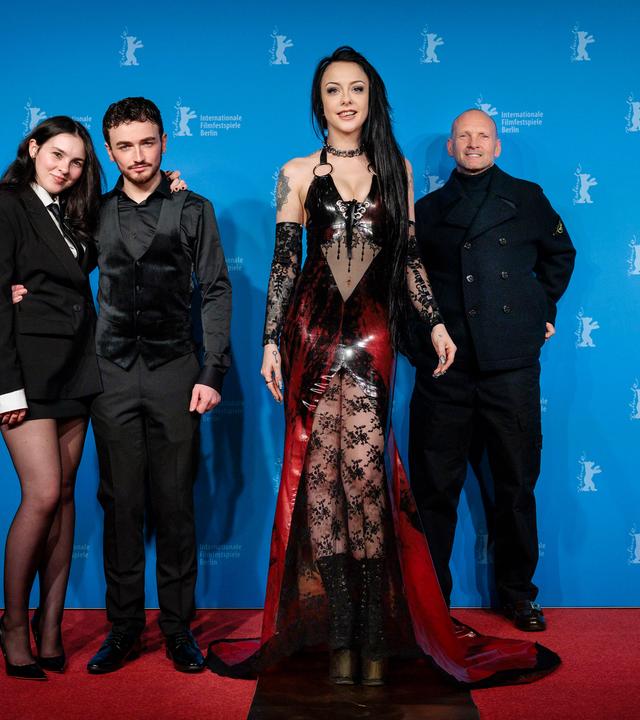 Vor dem blauen Hintergrund der Berlinale-Pressekonferenzen und auf einem roten Teppich stehen zwei Männer in schwarzer Kleidung und zwei Frauen. Eine junge Frau im Vordergrund hat lange schwarze Haare und trägt ein freizügiges Kostüm aus rotem Lack und schwarzer Spitze. Die zweite Frau trägt ein schwarzes Kostüm mit Minirock und hat den Arm um einen der Männer gelegt.