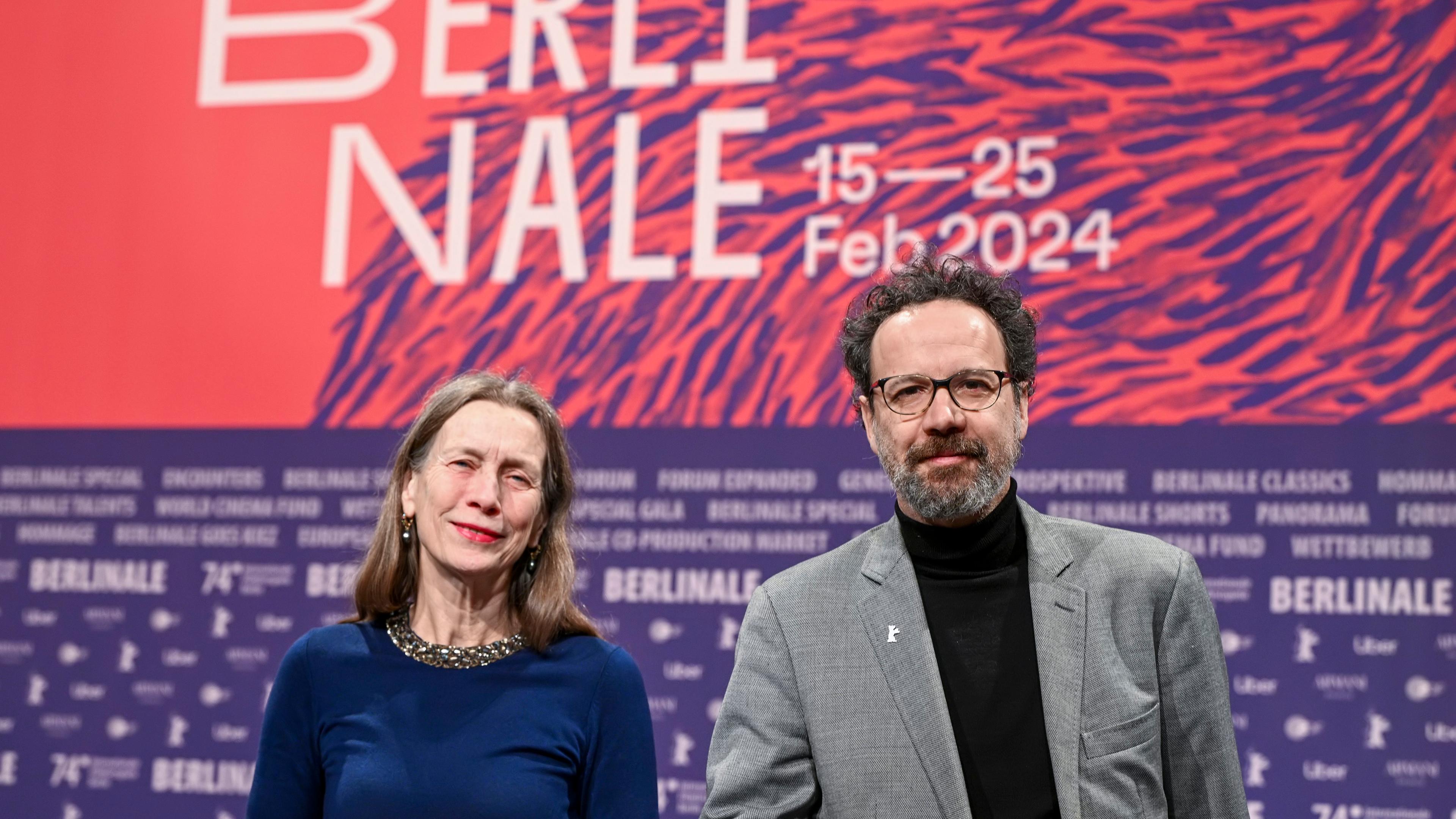Eine ältere Frau und ein Mann mit Bart stehen vor einem großen Plakat für die Berlinale 2024.