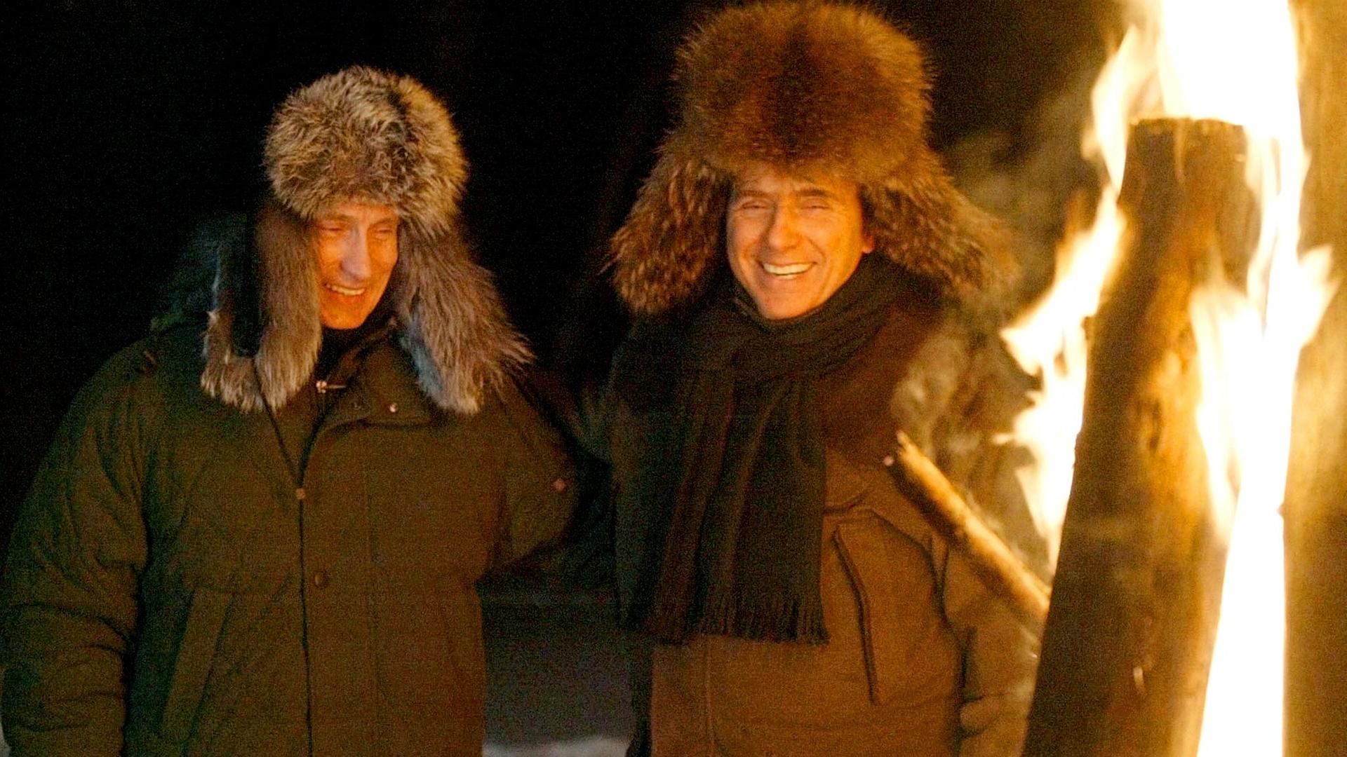 Putin und Berlusconi stehen mit warmer Kleidung und Pelzmützen an einem Feuer.