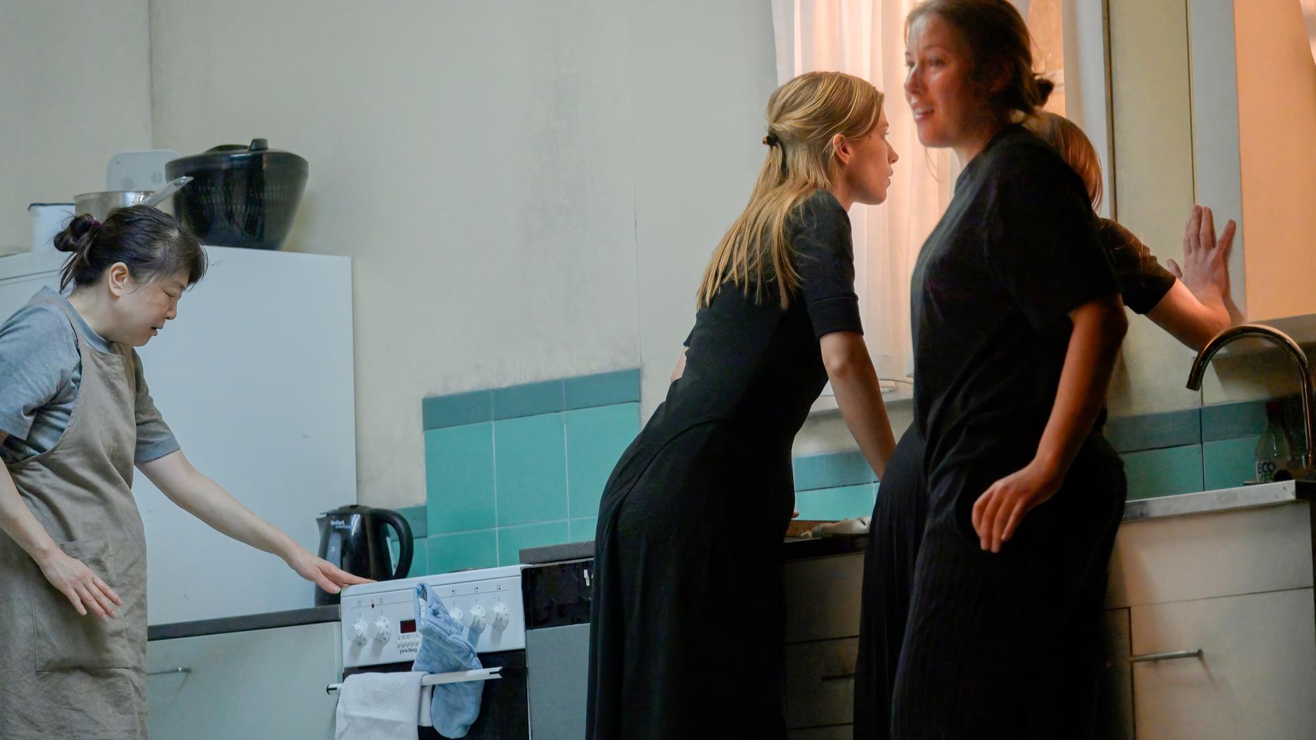 Vier Frauen in einer Küchenkulisse. Links bedient eine Frau in Schürze den Herd. In der Mitte beugt sich eine Frau in schwarzer Kleidung über die Spüle und schaut angespannt nach draußen. Neben ihr steht eine weitere Frau in Schwarz, die sich leicht zur Seite dreht und mit einem Lächeln den Raum betrachtet. Die vierte Frau steht dicht am Fenster und legt eine Hand an die Scheibe, während ihr Blick nach draußen geht. Die Szene wirkt angespannt und geheimnisvoll, geprägt von kühlen Blau- und Grautönen.