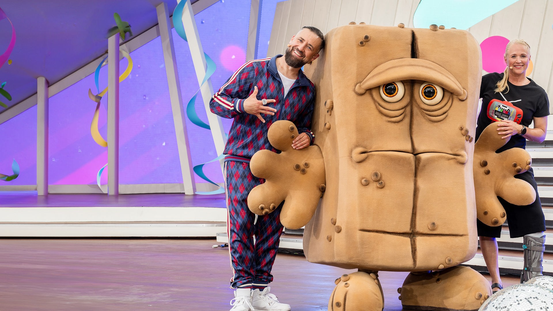 Moderator Bürger Lars Dietrich und Moderatorin Singa Gätgens stehen gut gelaunt in einem Studio. In der Mitte steht Bernd, das Brot und schaut schlecht gelaunt aus. 