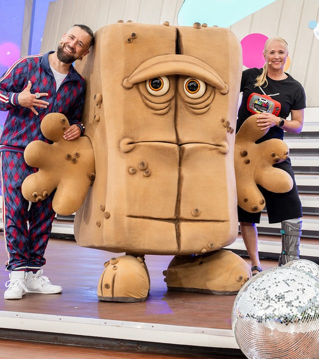 Moderator Bürger Lars Dietrich und Moderatorin Singa Gätgens stehen gut gelaunt in einem Studio. In der Mitte steht Bernd, das Brot und schaut schlecht gelaunt aus. 