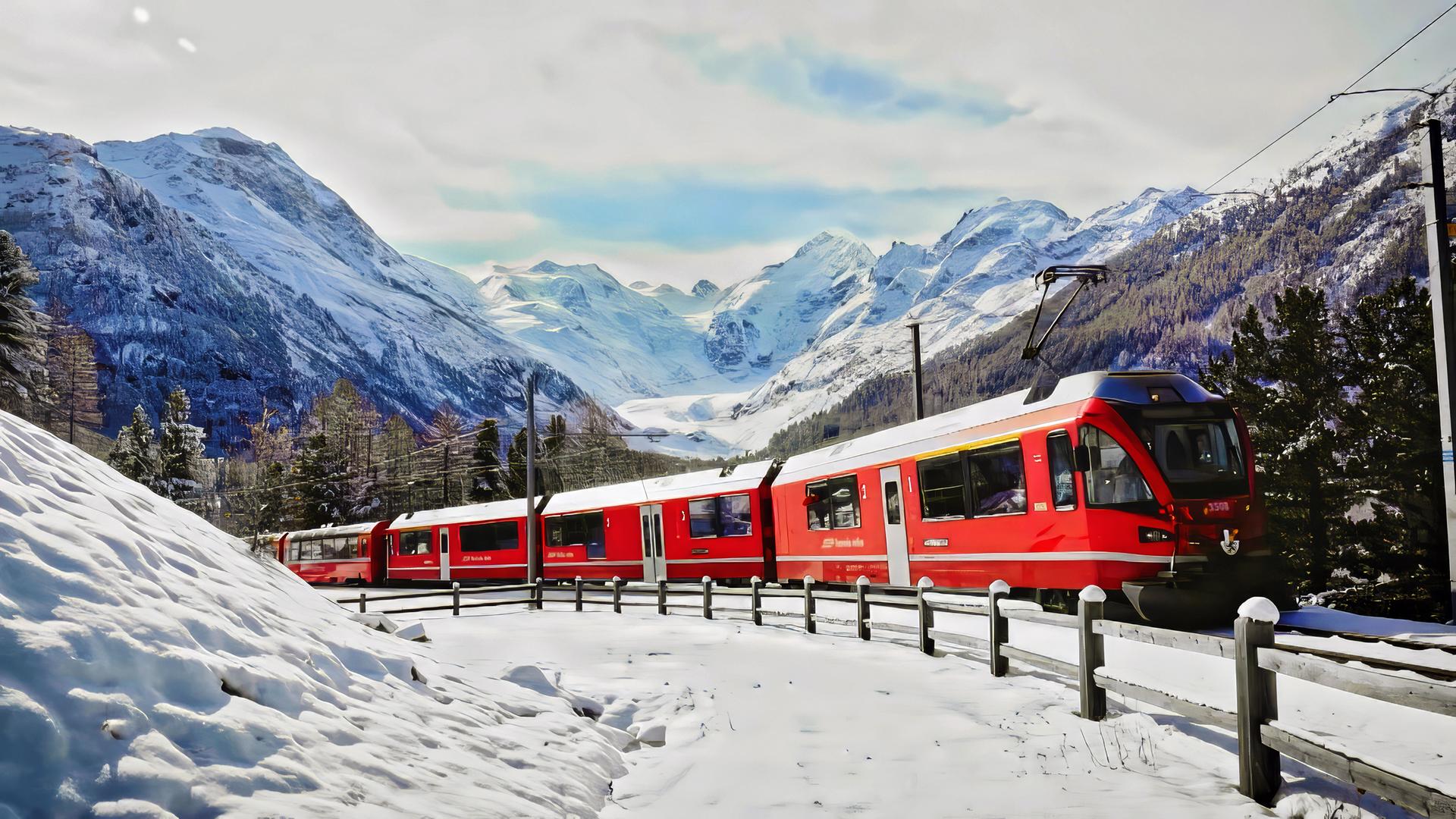 Eine rote Bahn fährt durch eine verschneite Gebirgslandschaft, umgeben von schneebedeckten Bäumen und hohen Bergen.