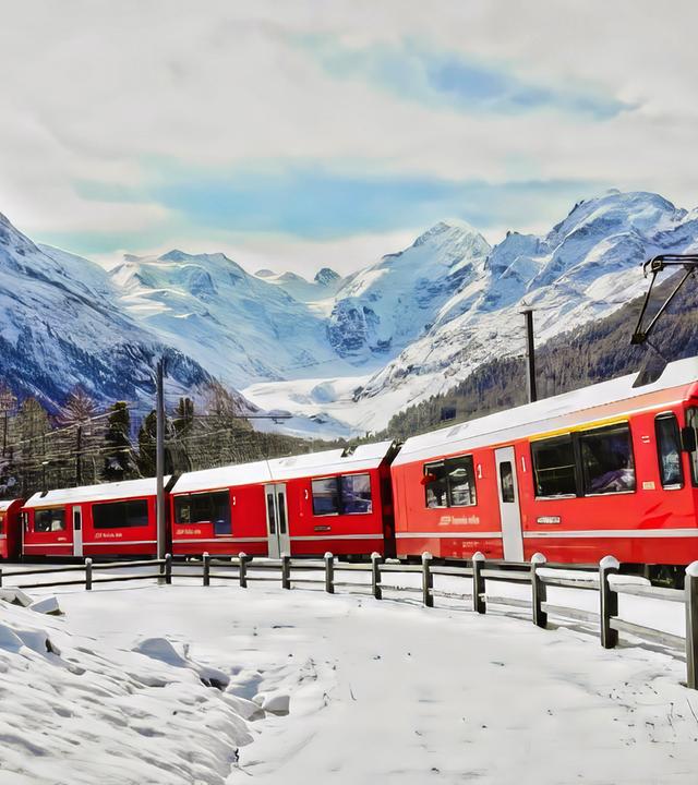 Eine rote Bahn fährt durch eine verschneite Gebirgslandschaft, umgeben von schneebedeckten Bäumen und hohen Bergen.