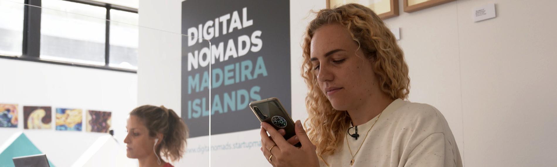 Zwei Frauen sitzen an einem Schreibtisch und arbeiten am Laptop. Die blonde Frau im Vordergrund, Merle Makobe, um die es geht, hält ein Handy in der Hand. Im Hintergrund auf der Wand steht geschrieben: Digital Nomads Madeira Islands. Übersetzung: Digitale Nomaden der Insel Madeira.