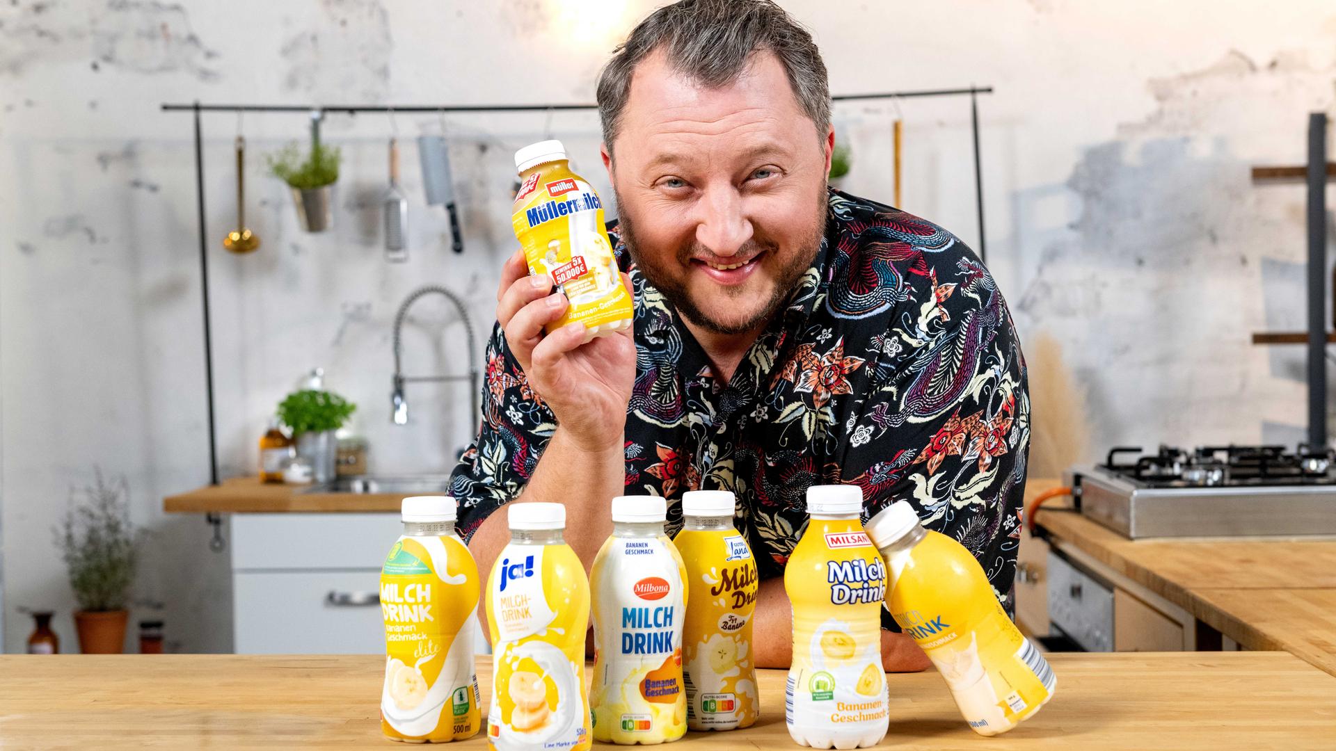 Sebastian Lege lehnt über einem Tisch und hält dabei einen Bananendrink in der Hand. Vor ihm stehen diverse Konkurrenzprodukte.