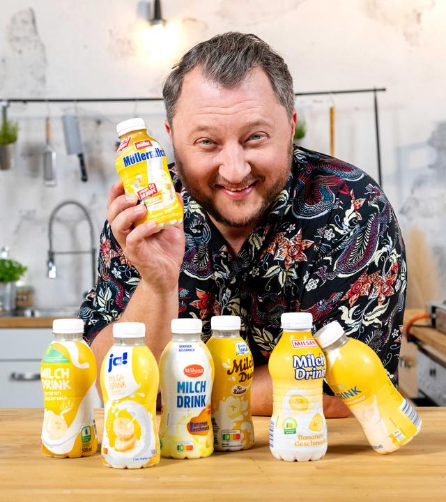 Sebastian Lege lehnt über einem Tisch und hält dabei einen Bananendrink in der Hand. Vor ihm stehen diverse Konkurrenzprodukte.