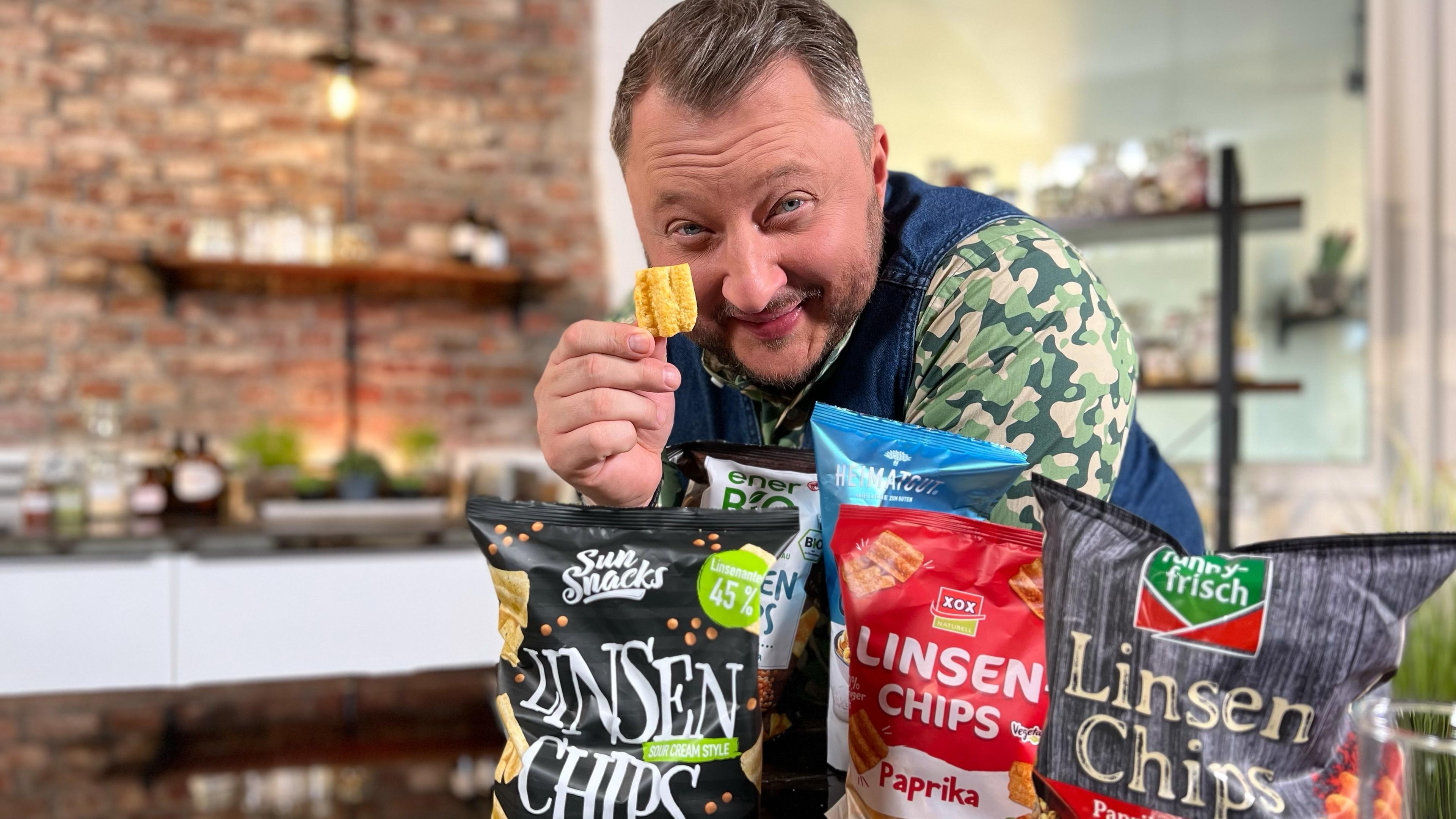 Sebastian Lege lehnt an einem Tisch, hält einen Linsenchip hoch. Vor ihm liegen diverse Linsenchips-Verpackungen.