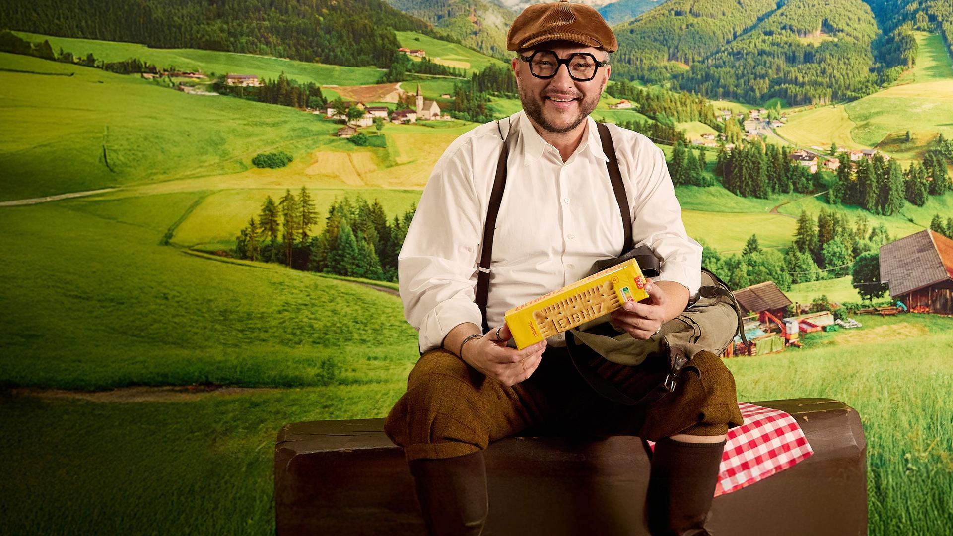 Sebastian Lege sitzt in traditionell aussehender Kleidung in einer Berglandschaftskulisse, in der Hand hält er eine Packung Kekse 