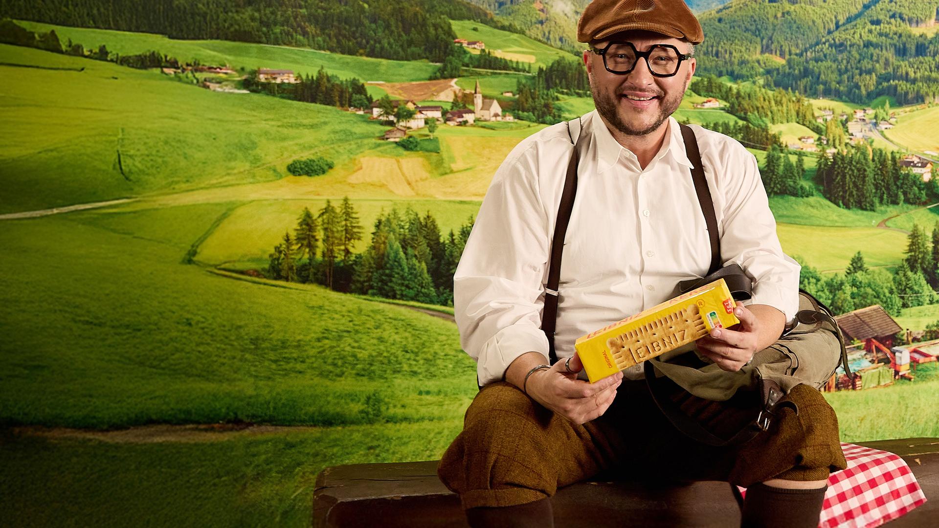 Sebastian Lege sitzt in traditionell aussehender Kleidung in einer Berglandschaftskulisse, in der Hand hält er eine Packung Kekse. 