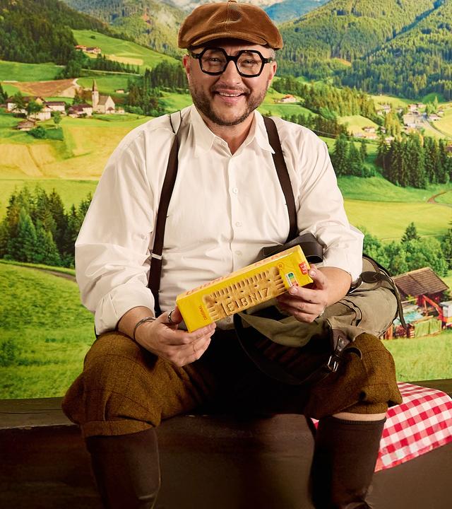 Sebastian Lege sitzt in traditionell aussehender Kleidung in einer Berglandschaftskulisse, in der Hand hält er eine Packung Kekse 