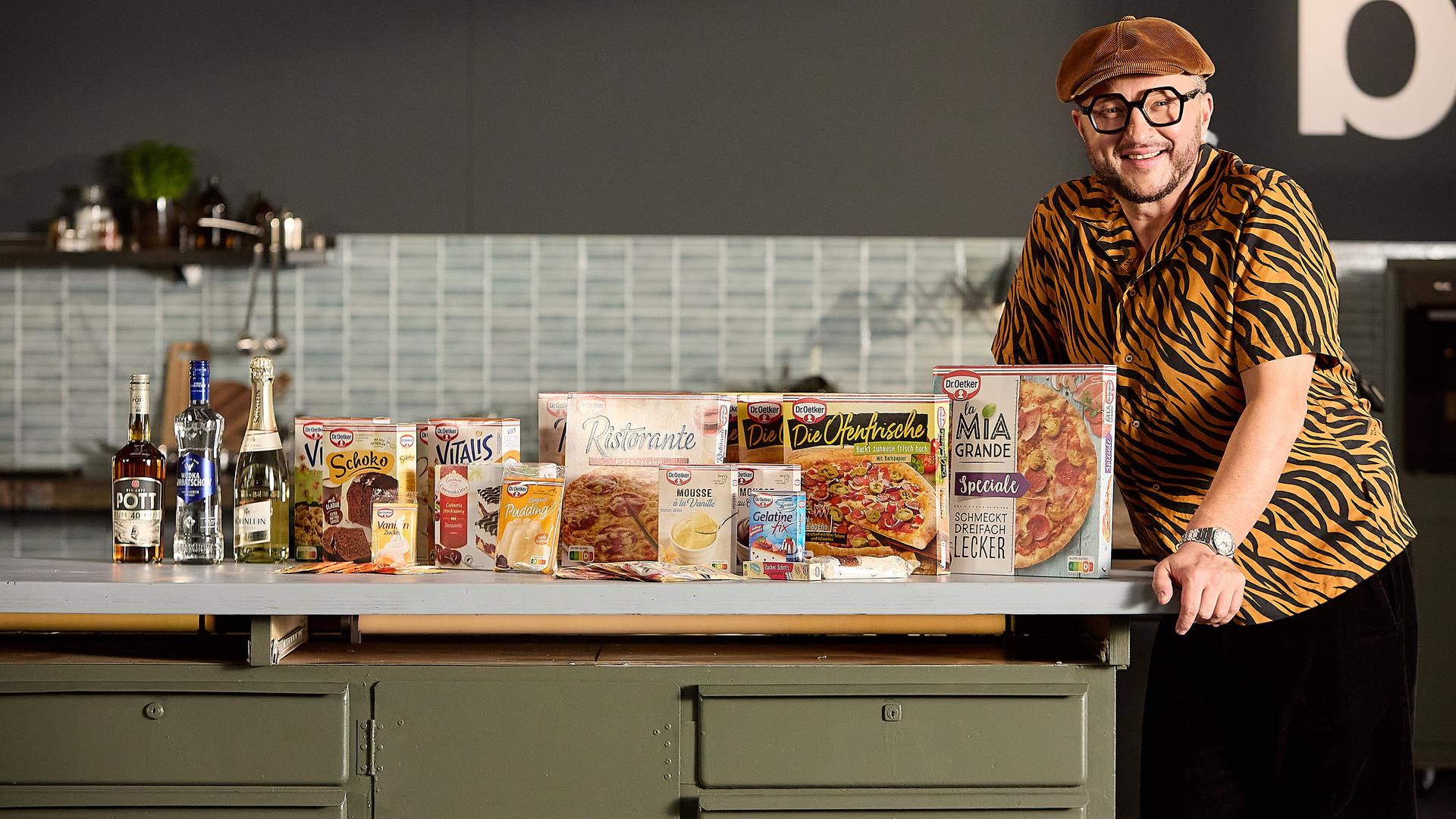 Sebastian steht am Tresen seiner Werkstattküche. Die Arbeitsfläche ist gefüllt mit Produkten von Dr. Oetker.