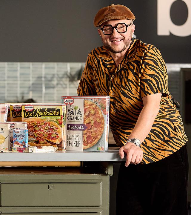 Sebastian steht am Tresen seiner Werkstattküche. Die Arbeitsfläche ist gefüllt mit Produkten von Dr. Oetker.