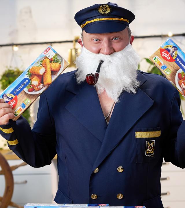 Sebastian Lege steht als Captain Iglo verkleidet und mit Pfeife im Mund vor einem Tisch mit mehreren Packungen Iglo Fischstäbchen.