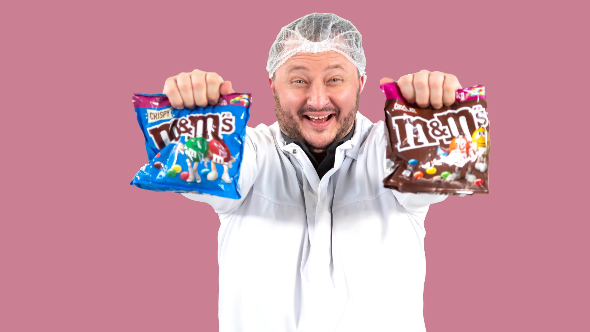 Sebastian Lege steht in einer Versuchsküche und hält zwei Packungen m&m's in den Händen.