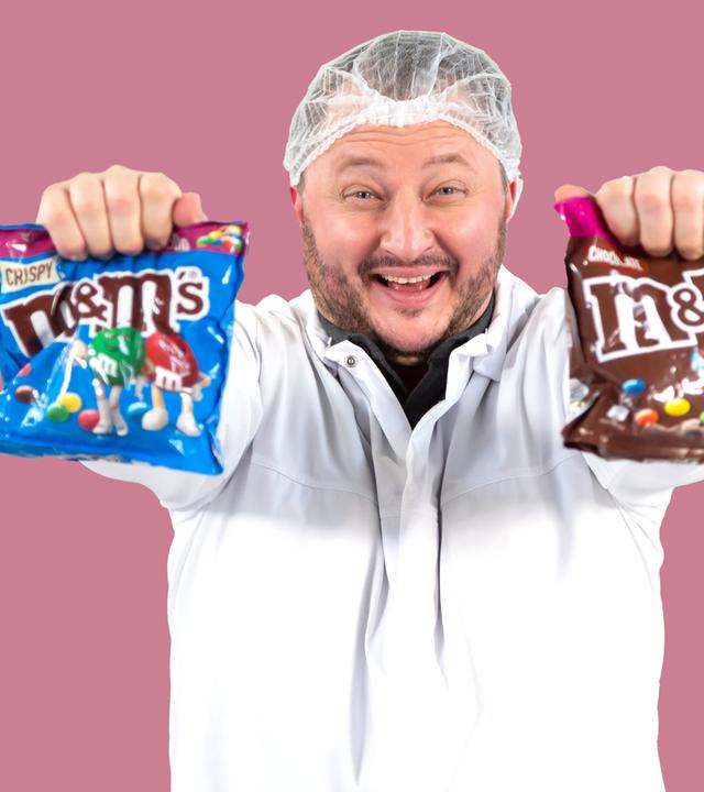 Sebastian Lege steht in einer Versuchsküche und hält zwei Packungen m&m's in den Händen.