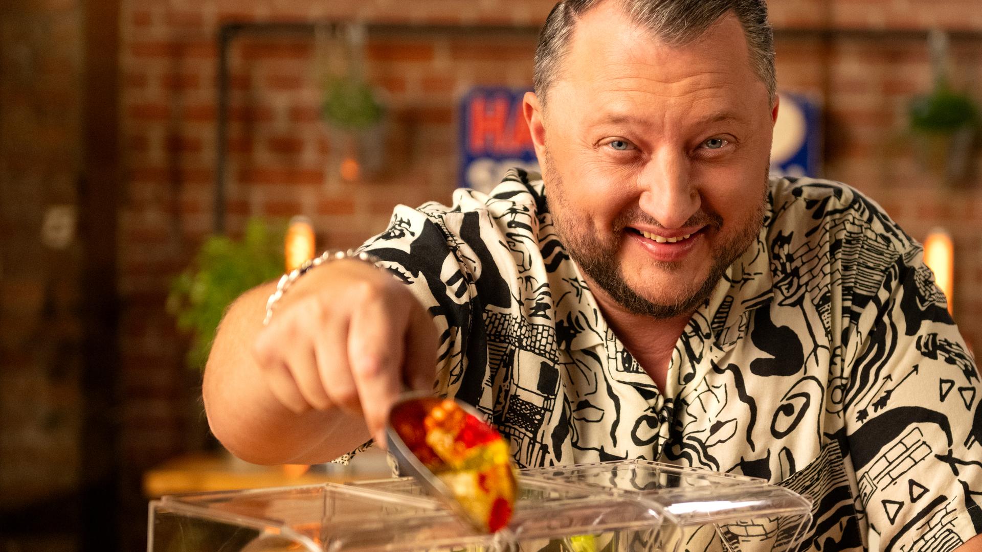 Sebastian Lege steht in seiner Küche hinter einer Süßigkeiten-Theke. In seiner Hand hält er eine Schaufel voll mit Goldbären.