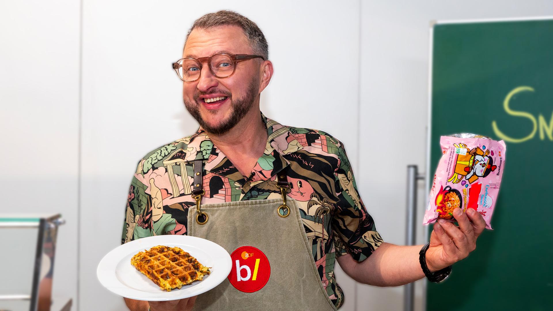 besseresser Kids - Lege packt´s an: Sebastian Lege steht in einem Klassenzimmer und hält in der einen Hand einen Teller, auf dem einen Waffel liegt. In der anderen Hand hält er eine Packung Instantnudeln.