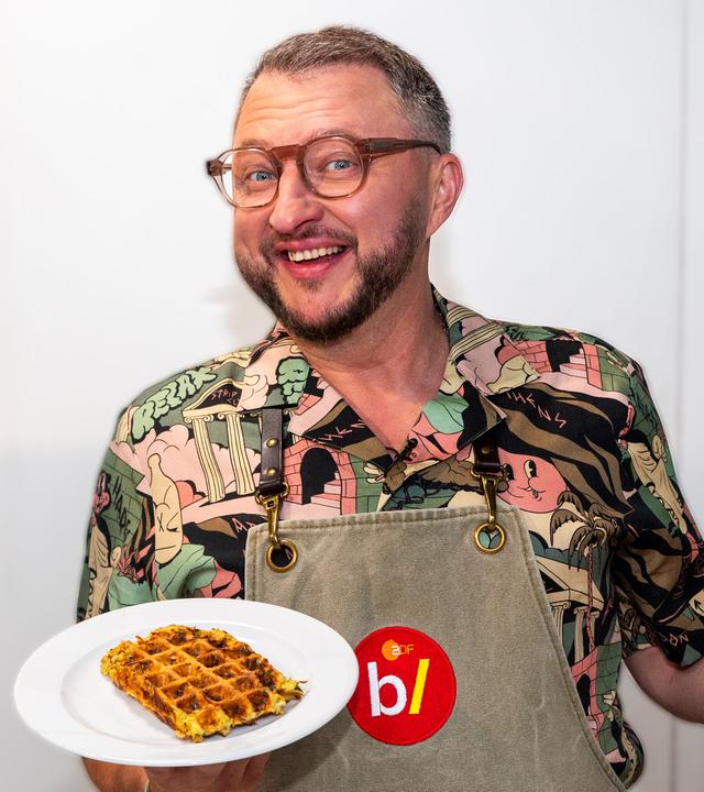 besseresser Kids - Lege packt´s an: Sebastian Lege steht in einem Klassenzimmer und hält in der einen Hand einen Teller, auf dem einen Waffel liegt. In der anderen Hand hält er eine Packung Instantnudeln.