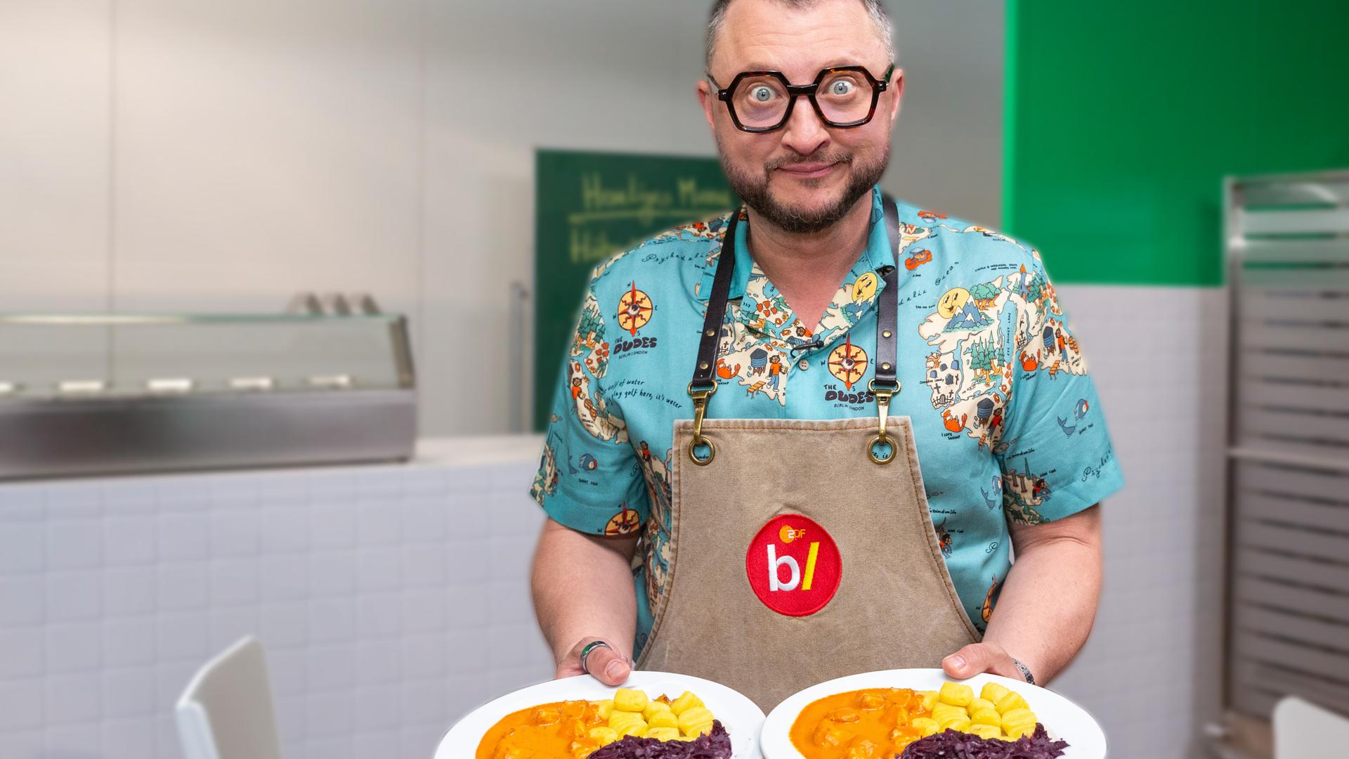 besseresser Kids - Lege packt´s an: Sebastian Lege steht in einem Klassenraum und hält in jeder Hand einen Teller mit einem Gericht aus Gnocchi, Rotkohl und Gulasch.