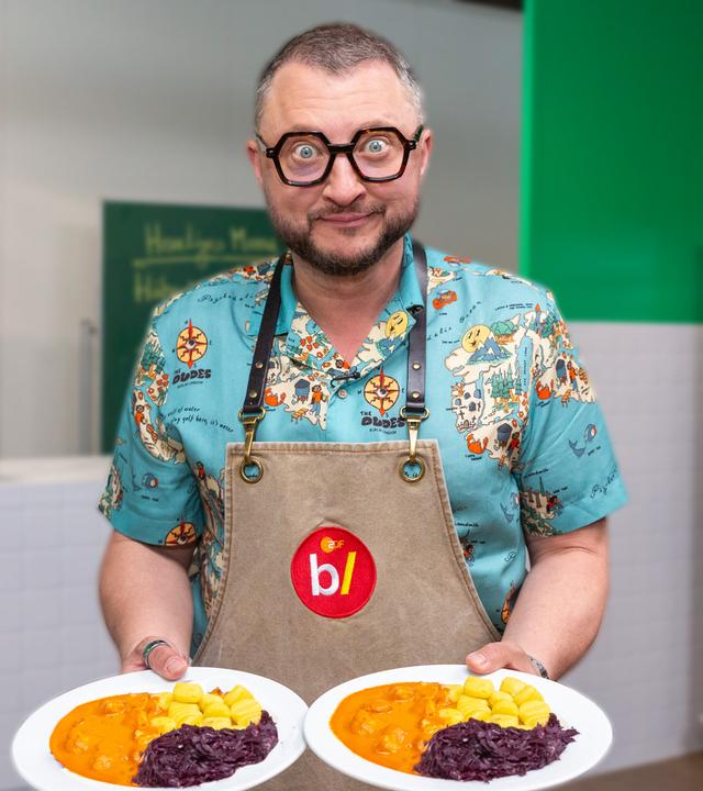 besseresser Kids - Lege packt´s an: Sebastian Lege steht in einem Klassenraum und hält in jeder Hand einen Teller mit einem Gericht aus Gnocchi, Rotkohl und Gulasch.