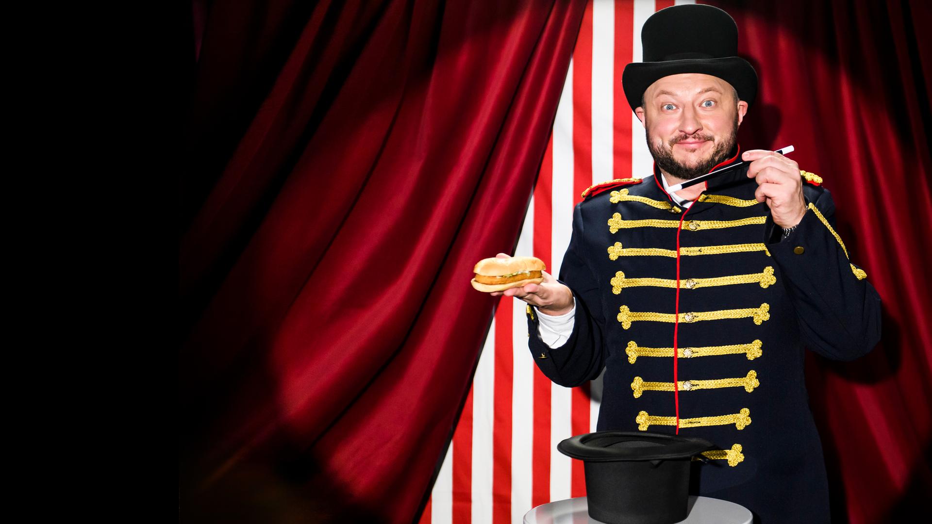 Sebastian Lege steht in einem Zaubererkostüm vor einem Zirkusvorhang und hält einen Burger und einen Zauberstab in der Hand.