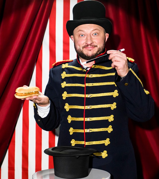 Sebastian Lege steht in einem Zaubererkostüm vor einem Zirkusvorhang und hält einen Burger und einen Zauberstab in der Hand.