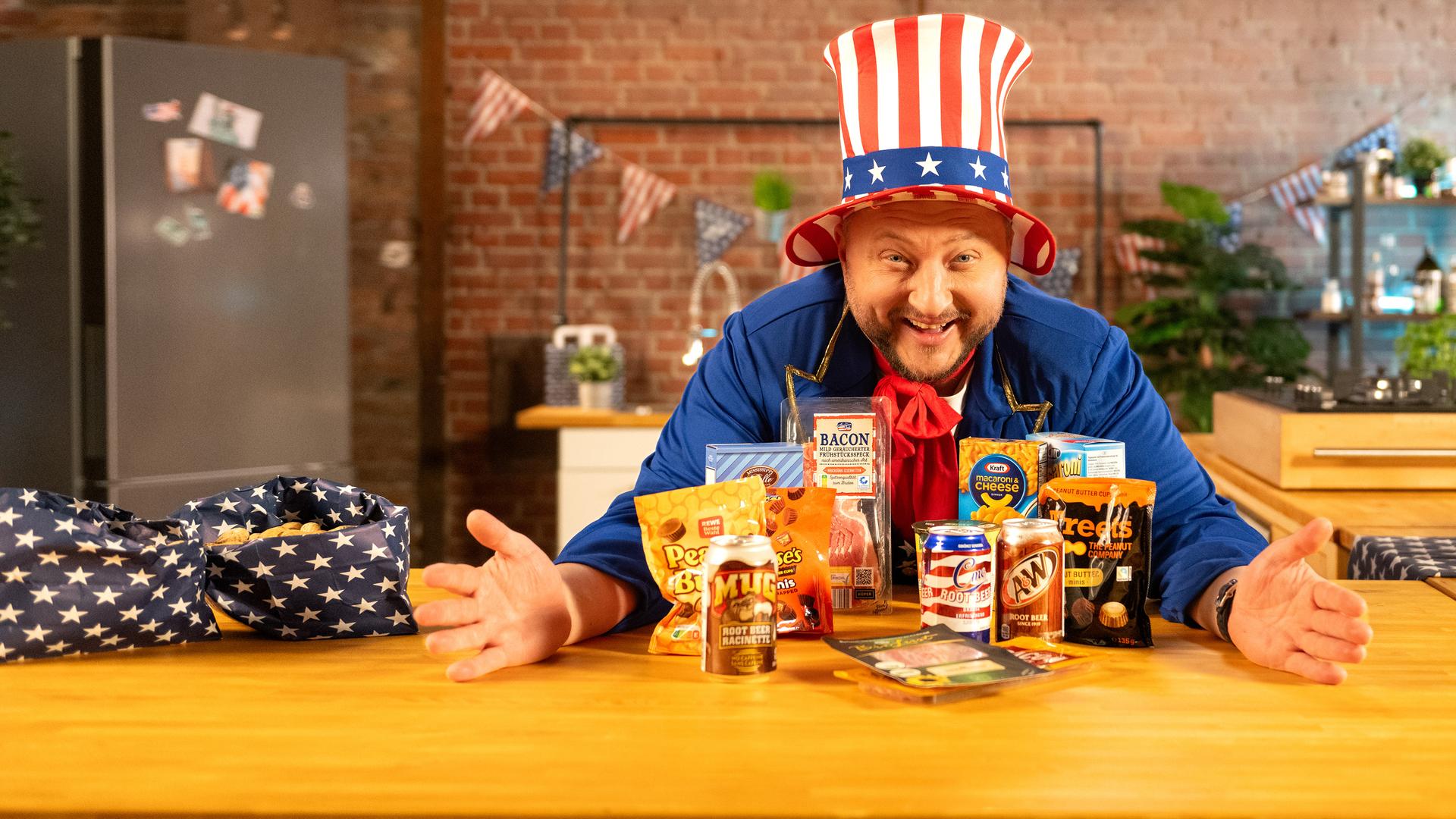 Sebastian Lege steht lachend hinter einer Küchentheke, auf der verschiedene Snacks aus den USA stehen. Er trägt einen Hut in den Farben der US-Flagge.